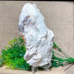 Pedra de Larimar em bruto com textura branca e detalhes azuis, apoiada em suporte metálico sobre fundo neutro, com folhagem decorativa ao lado.