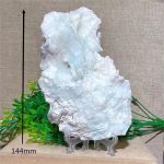 Pedra de larimar em bruto de grande dimensão, com textura irregular e cor azul clara, apoiada em suporte de acrílico transparente, com plantas decorativas ao fundo.