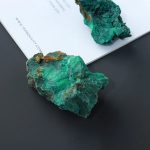 Malaquite bruta de cor verde com detalhes marrons, vista de perto sobre fundo escuro e uma folha de papel ao fundo.