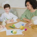 Criança e adulta a montar uma maquete do sistema solar com bolas coloridas representando planetas, numa mesa de sala de aula ou estudo.