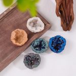 Conjunto de cinco geodos de cristal de rocha com diferentes cores e texturas, alguns com brilho e outros com cristais visíveis, sobre uma superfície branca com decoração de madeira e planta.