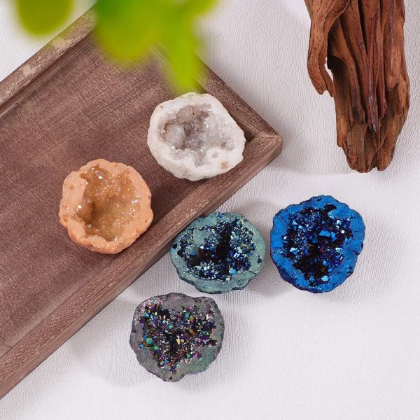 Conjunto de cinco geodos de cristal de rocha com diferentes cores e texturas, alguns com brilho e outros com cristais visíveis, sobre uma superfície branca com decoração de madeira e planta.