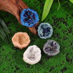 Conjunto de cinco mini geodos de cristal de rocha em cores variadas, exibidos sobre relva verde com folhas ao redor, destacando detalhes internos e texturas.