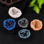 Conjunto de cinco mini geodos de cristal de rocha com diferentes cores e texturas, exibidos sobre uma superfície escura com uma planta ao fundo.