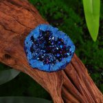 Geodo de cristal de rocha cinzento com interior brilhante e fragmentos azuis, colocado sobre tronco de madeira com fundo de plantas verdes.