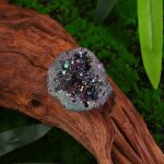 Geodo de cristal de rocha cinzento com cristais iridescentes, colocado sobre tronco de madeira com vegetação verde ao fundo.