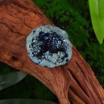 Mini geodo de cristal de rocha cinzento com cristais azuis e prateados, colocado numa peça de madeira, com fundo de vegetação verde.