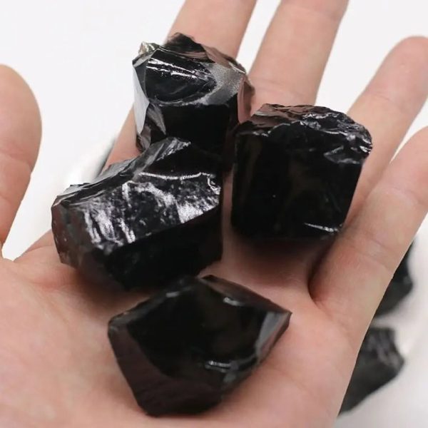 Quatro pedaços de obsidiana negra com superfície brilhante, exibidos na palma da mão, mostrando diferentes ângulos e texturas.