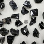 Amostras de obsidiana em bruto com superfícies brilhantes e formas irregulares, dispostas sobre uma superfície clara, com uma moeda de um euro para escala.