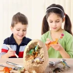 Crianças explorando um ovo de dinossauro para escavar, com dinossauros de plástico ao redor e um ovo parcialmente aberto na mesa.