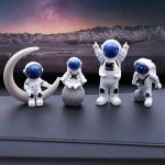 Quatro estatuetas de astronautas com capacetes azuis, uma delas com braços levantados, colocadas sobre rochas simuladas com uma paisagem montanhosa ao fundo.