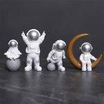 Quatro estatuetas de astronautas em diferentes poses, uma com uma lua de fundo, todas com capacetes prateados e trajes brancos, em fundo escuro.