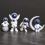 Quatro estatuetas de astronautas com capacetes azuis decorados com estrelas, cada uma em diferentes posições e tamanhos, com uma delas sentada numa rocha, outra com braços levantados, e uma com uma lu.