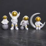 Conjunto de quatro estatuetas de astronautas com capacetes dourados, um sentado na lua, outro de pé com braços levantados, um de lado na superfície lunar e um na lua com uma lua crescente ao lado.