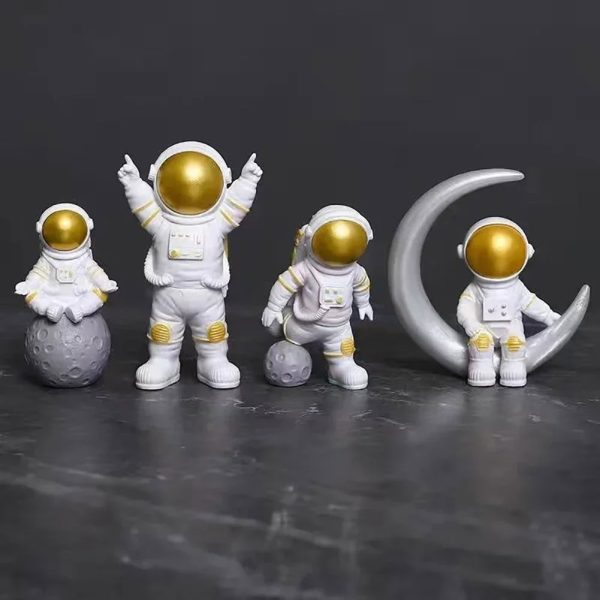 Conjunto de quatro estatuetas de astronautas com capacetes dourados, um sentado na lua, outro de pé com braços levantados, um de lado na superfície lunar e um na lua com uma lua crescente ao lado.