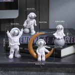 Quatro estatuetas de astronautas em diferentes poses, uma com uma lua de suporte, uma com uma bola, uma com uma mochila e uma com uma bola de plasma, todas em branco com detalhes dourados.