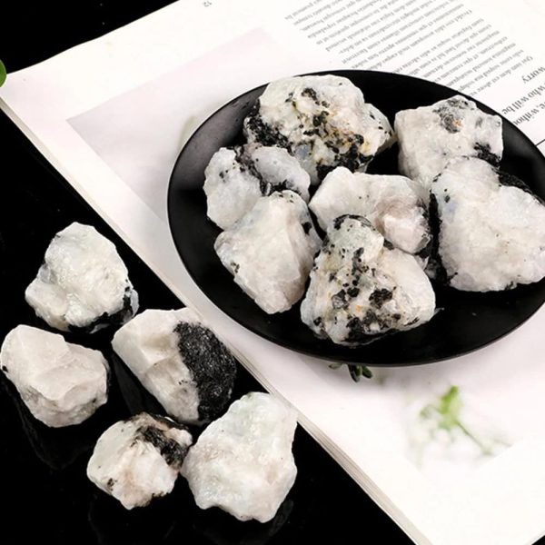 Pedras da Lua brutas com superfície irregular, algumas com manchas negras, dispostas numa tigela preta e espalhadas numa superfície branca.