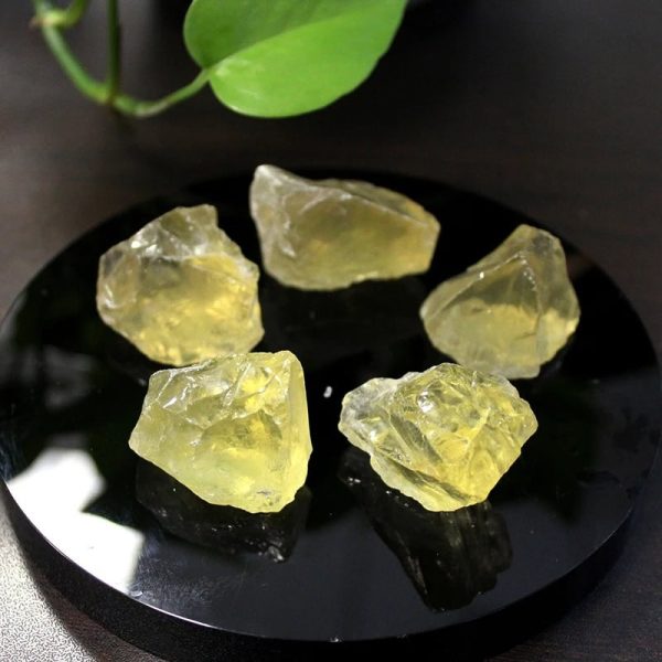 Seis pedaços de citrino bruto de cor amarela, com superfícies irregulares e translucidez, dispostos sobre uma base preta, com uma planta ao fundo.