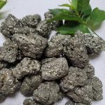 Conjunto de pedras de pirite em bruto com superfície brilhante e textura granular, acompanhadas de folhas verdes de planta decorativa.