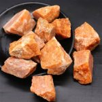 Vários pedaços de pedra do sol em bruto, com tonalidade laranja e textura áspera, dispostos numa tigela preta.
