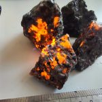 Quatro pedras Yooperlite com fluorescência laranja brilhante sob luz UV, destacando suas texturas e cores escuras, colocadas sobre uma superfície branca com régua para escala.