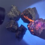 Grupo de pedras Yooperlite com fluorescência laranja e roxa sob luz UV, destacando-se contra fundo escuro.