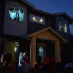 Projeções assustadoras de olhos, fantasmas e esqueletos nas janelas de uma casa à noite, com pessoas assistindo na rua.