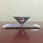 Projeção de um coração em luz rosa a partir de um projetor holográfico colocado sobre um telemóvel branco, com fundo neutro.