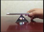 Dispositivo de projeção holográfica em forma de pirâmide com telemóvel apoiado por cima, projetando uma imagem tridimensional.