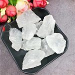 Vários cristais de quartzo branco em bruto, com superfícies irregulares e translúcidas, dispostos numa tigela preta sobre uma superfície cinzenta, com flores ao fundo.