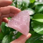 Pedra de quartzo rosa bruta, com superfície irregular e translúcida, segurada por uma mão, com folhas verdes ao fundo.