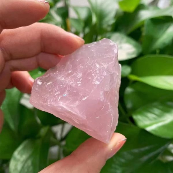 Pedra de quartzo rosa bruta, com superfície irregular e translúcida, segurada por uma mão, com folhas verdes ao fundo.