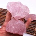 Dois cristais de quartzo rosa em bruto, com superfícies irregulares e cor rosa pálido, sendo segurados por uma mão, com fundo de esteira de bambu.
