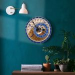 Relógio astronómico de parede com detalhes em azul e dourado, mostrando fases lunares e constelações, instalado numa parede verde escura.