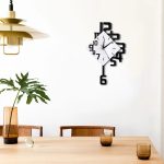 Relógio de parede com números grandes e ponteiros pretos, com forma de quadrado e círculo, decorando uma parede branca.
