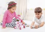 Duas crianças brincam com um robô rosa com olhos grandes e rodas, numa sala bem iluminada, com brinquedos ao fundo.