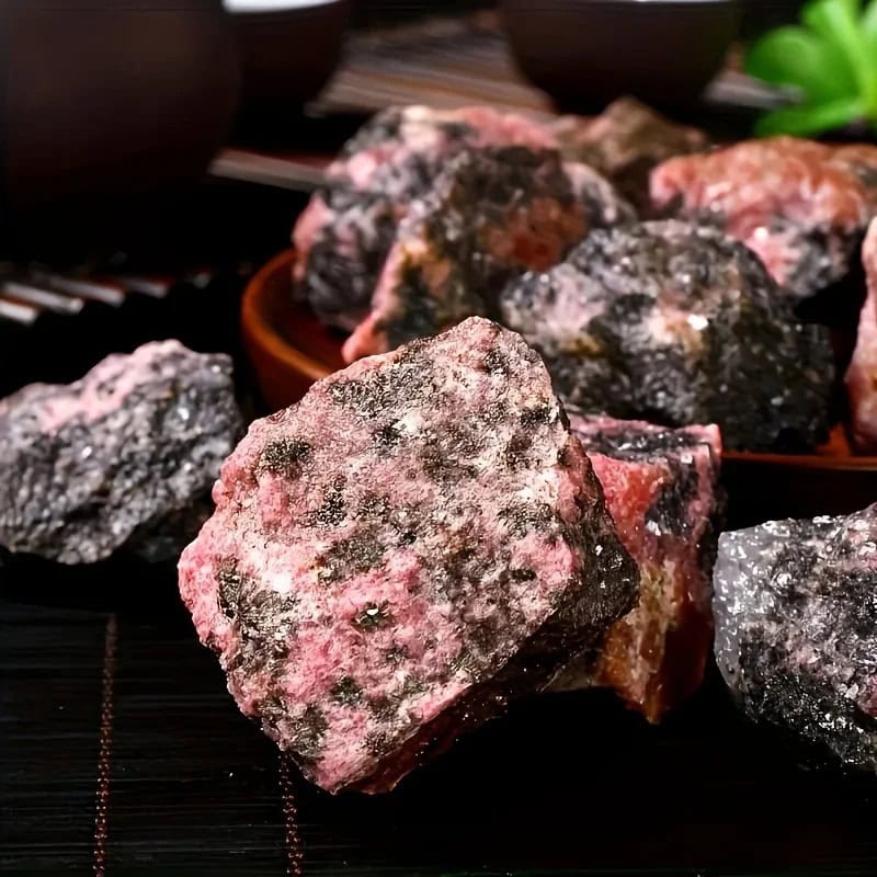 Pedras de rodonita bruta com cores rosadas, pretas e brancas, textura irregular e superfície áspera, exibidas numa bandeja de madeira.
