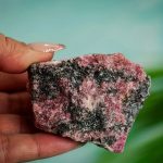 Pedra de rodoíta com cores rosa, preto e cinza, mostrando textura irregular e tamanho médio, segurada por uma mão com unhas bem cuidadas.