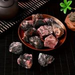 Rodeonita em bruto com cores variadas, incluindo rosa, preto e cinza, exibida numa tigela de barro escura, com detalhes de textura e forma irregular.