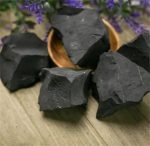 Pedaços de shungite em bruto de forma irregular, com superfície escura e textura rugosa, dispostos numa tigela de madeira sobre uma mesa de madeira, com flores roxas ao fundo.