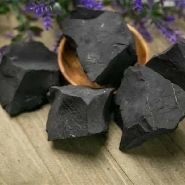 Pedaços de shungite em bruto de forma irregular, com superfície escura e textura rugosa, dispostos numa tigela de madeira sobre uma mesa de madeira, com flores roxas ao fundo.