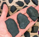Três pedaços de shungite bruta de diferentes tamanhos na palma da mão, com mais pedaços espalhados ao fundo sobre uma superfície de madeira.