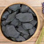 Pedras de shungite brutas de cor escura, com formas irregulares, colocadas numa tigela de madeira sobre uma superfície de madeira clara.