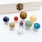 Conjunto de oito pedras preciosas de diferentes cores e tamanhos, cada uma em suporte de prata, com uma caixa de madeira ao fundo.