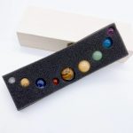 Conjunto de oito pedras preciosas representando os planetas do Sistema Solar, cada uma com cores distintas e tamanhos variados, colocadas numa espuma preta.