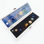 Conjunto de nove pedras preciosas representando os planetas do sistema solar, cada uma com cores distintas, dentro de uma caixa com espuma preta.