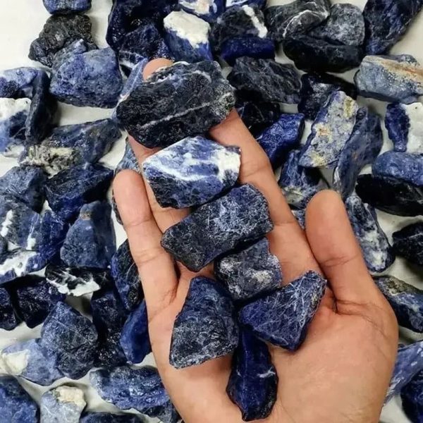 Mão segurando várias pedras de sodalita bruta de diferentes tamanhos e formas, com fundo de mais pedras espalhadas.