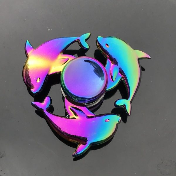Três delfins de metal iridescente com cores vibrantes, dispostos em círculo ao redor de um objeto circular central, sobre uma superfície escura.