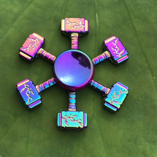 Spinner de mão com seis braços, cada um com detalhes em cores iridescentes e símbolos de trovão, colocado sobre uma folha verde.