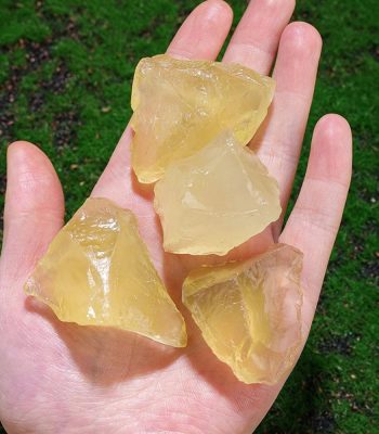 Quatro pedaços de topázio amarelo bruto na palma da mão, com fundo de relva verde.
